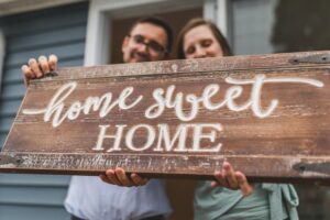 Smiling new homeowners holding a rustic wooden sign that reads ‘home sweet home’ after closing a property purchase directly from the seller.